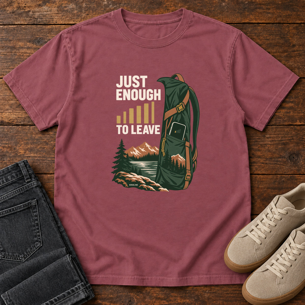 Brick Just Enough Signal to Leave T-Shirt with a green hiking backpack, mountains and gold signal bars beside cream lettering, front flat lay on wood.