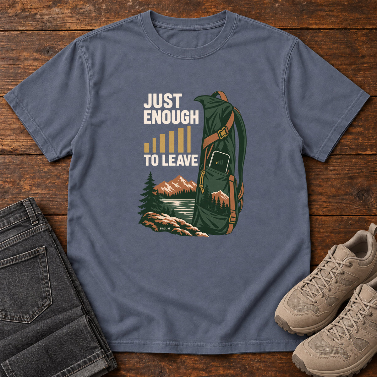 Denim Just Enough Signal to Leave T-Shirt with a green hiking backpack, mountains and gold signal bars beside cream lettering, front flat lay on wood.