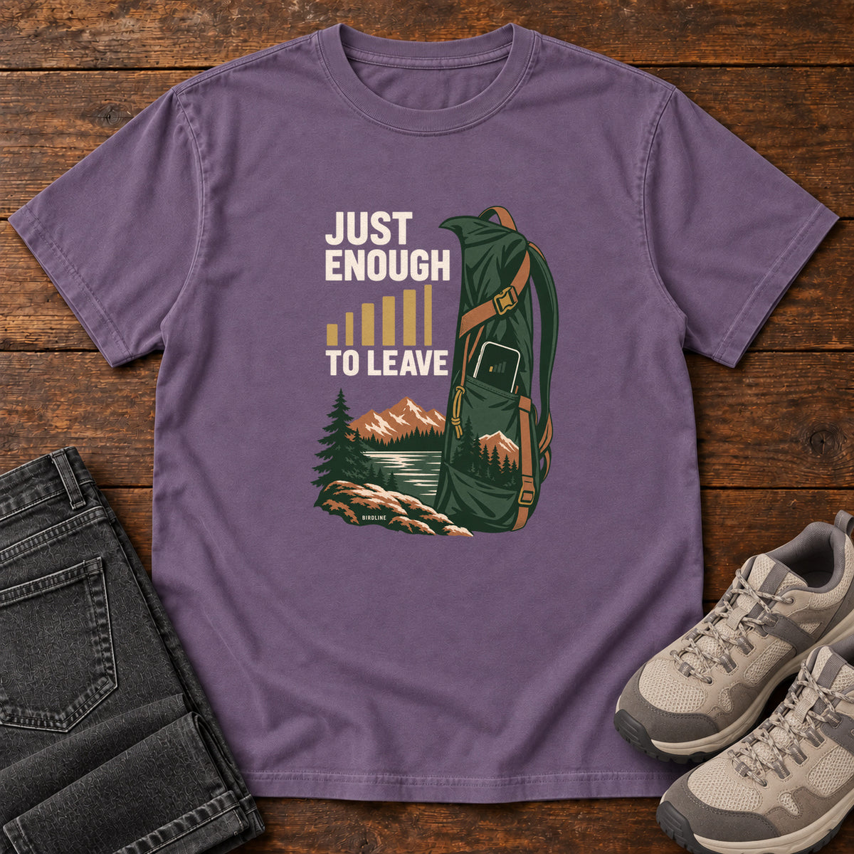 Grape Just Enough Signal to Leave T-Shirt with a green hiking backpack, mountains and gold signal bars beside cream lettering, front flat lay on wood.