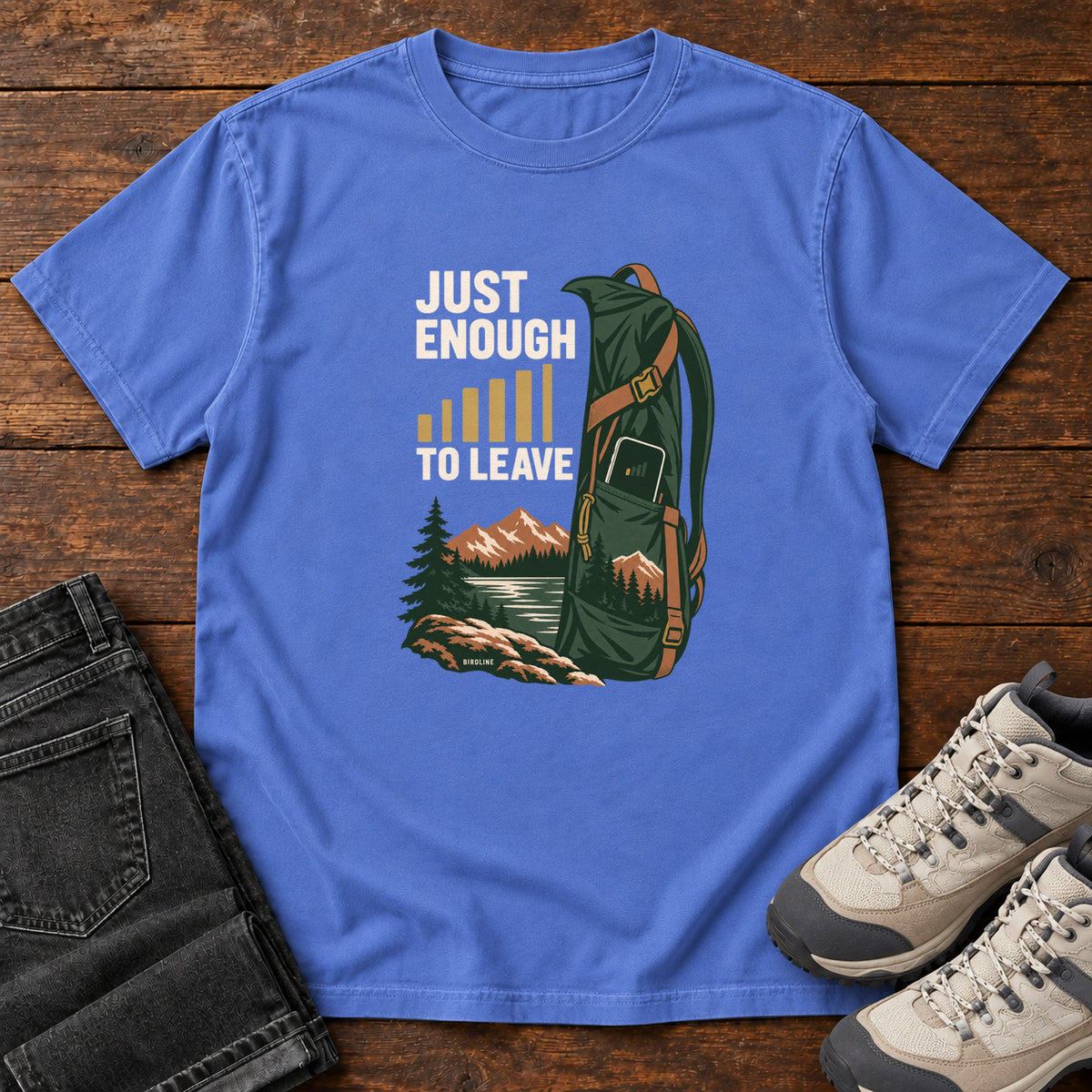 Mystic Blue Just Enough Signal to Leave T-Shirt with a green hiking backpack, mountains and gold signal bars beside cream lettering, front flat lay on wood.