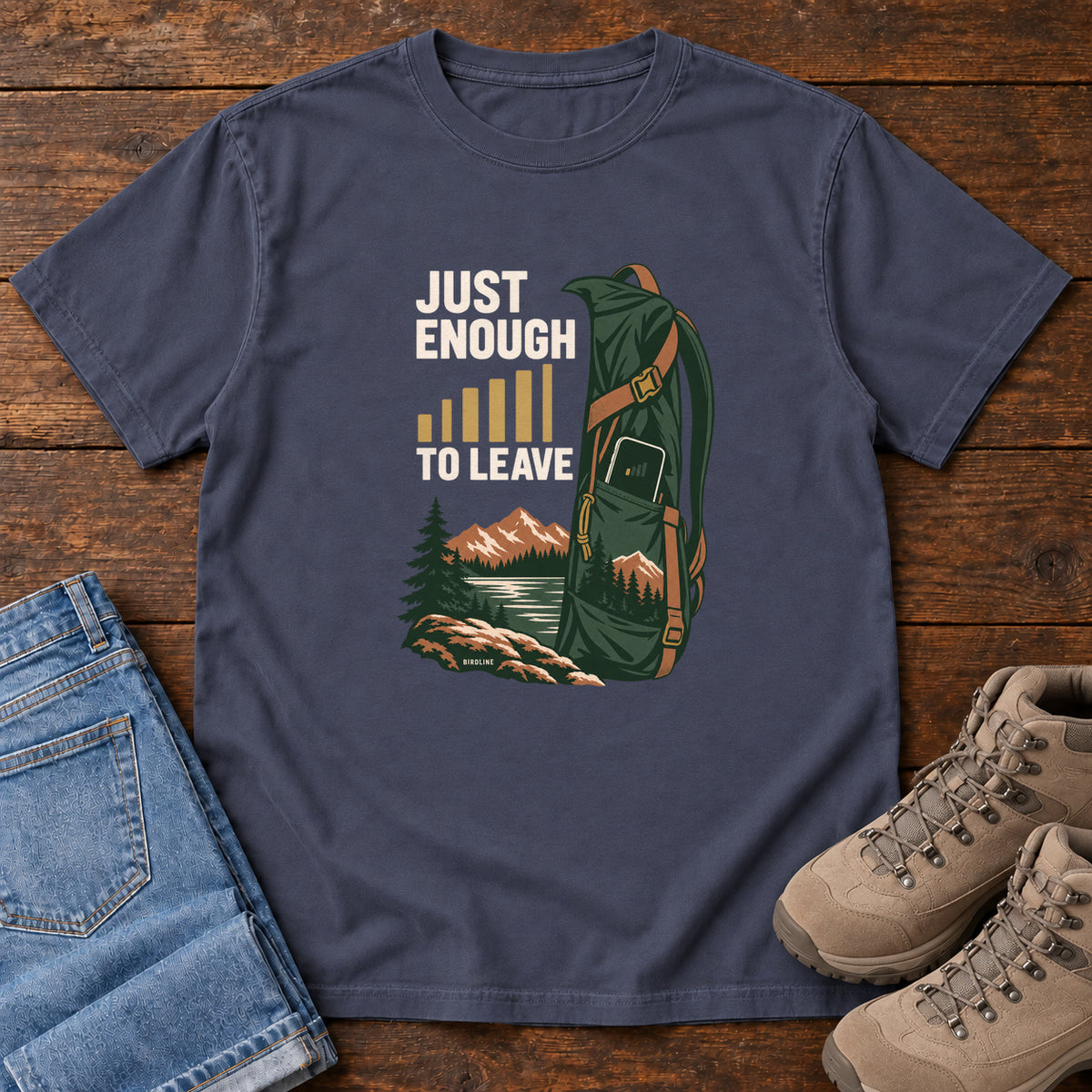 Navy Just Enough Signal to Leave T-Shirt with a green hiking backpack, mountains and gold signal bars beside cream lettering, front flat lay on wood.
