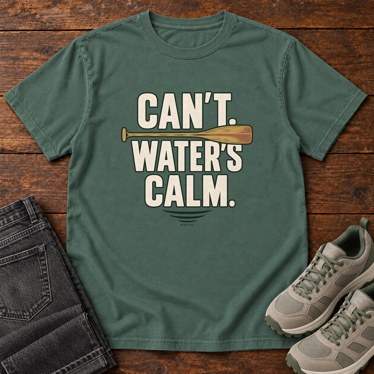 Can't. Water's Calm. T-Shirt in Blue Spruce, front laid flat on wood, featuring bold cream slogan lettering divided by a horizontal orange paddle.