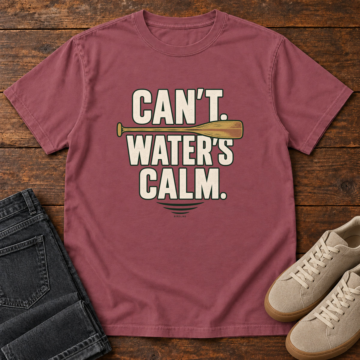 Can't. Water's Calm. T-Shirt in Brick, front laid flat on wood, featuring bold cream slogan lettering divided by a horizontal orange paddle.