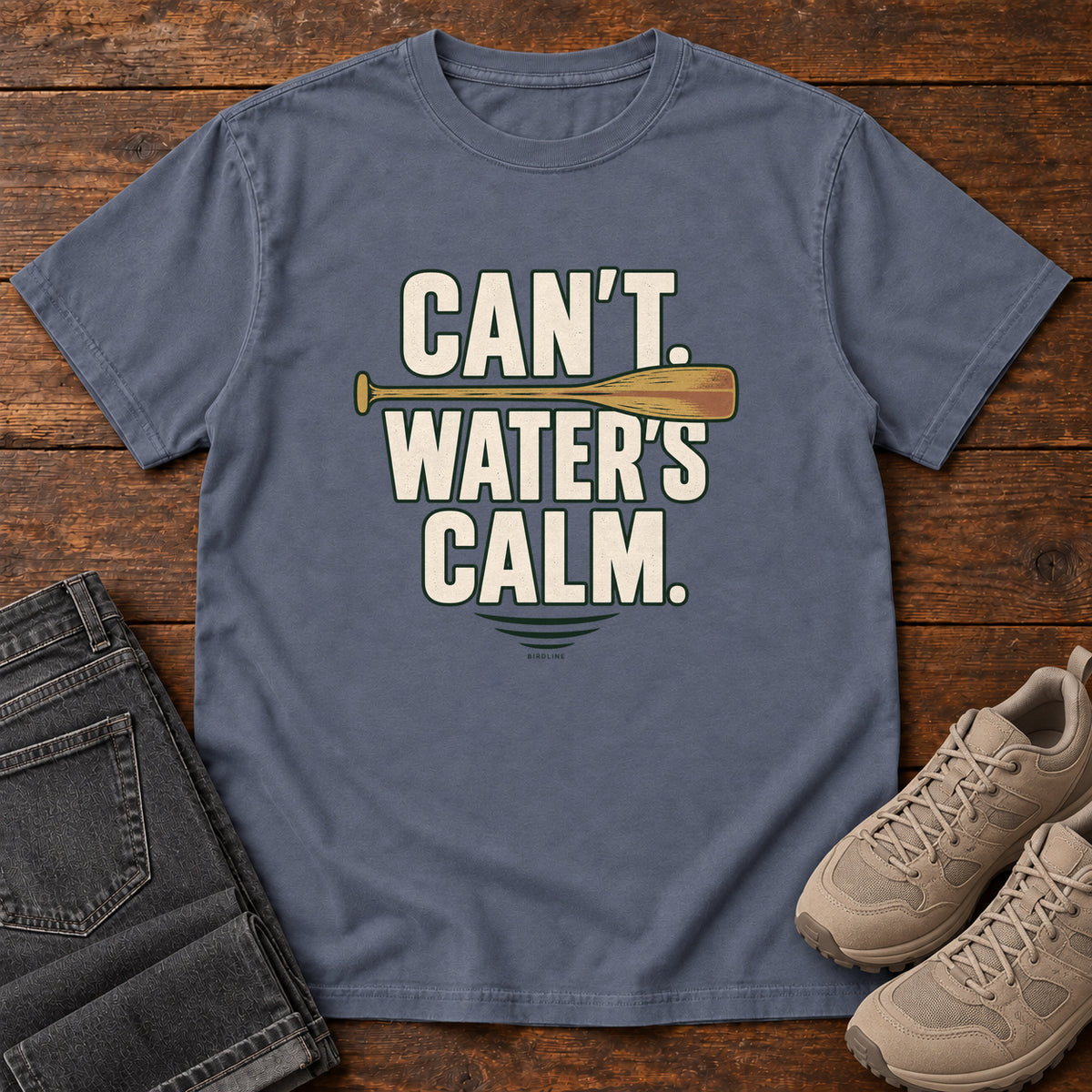 Can't. Water's Calm. T-Shirt in Denim, front laid flat on wood, featuring bold cream slogan lettering divided by a horizontal orange paddle.