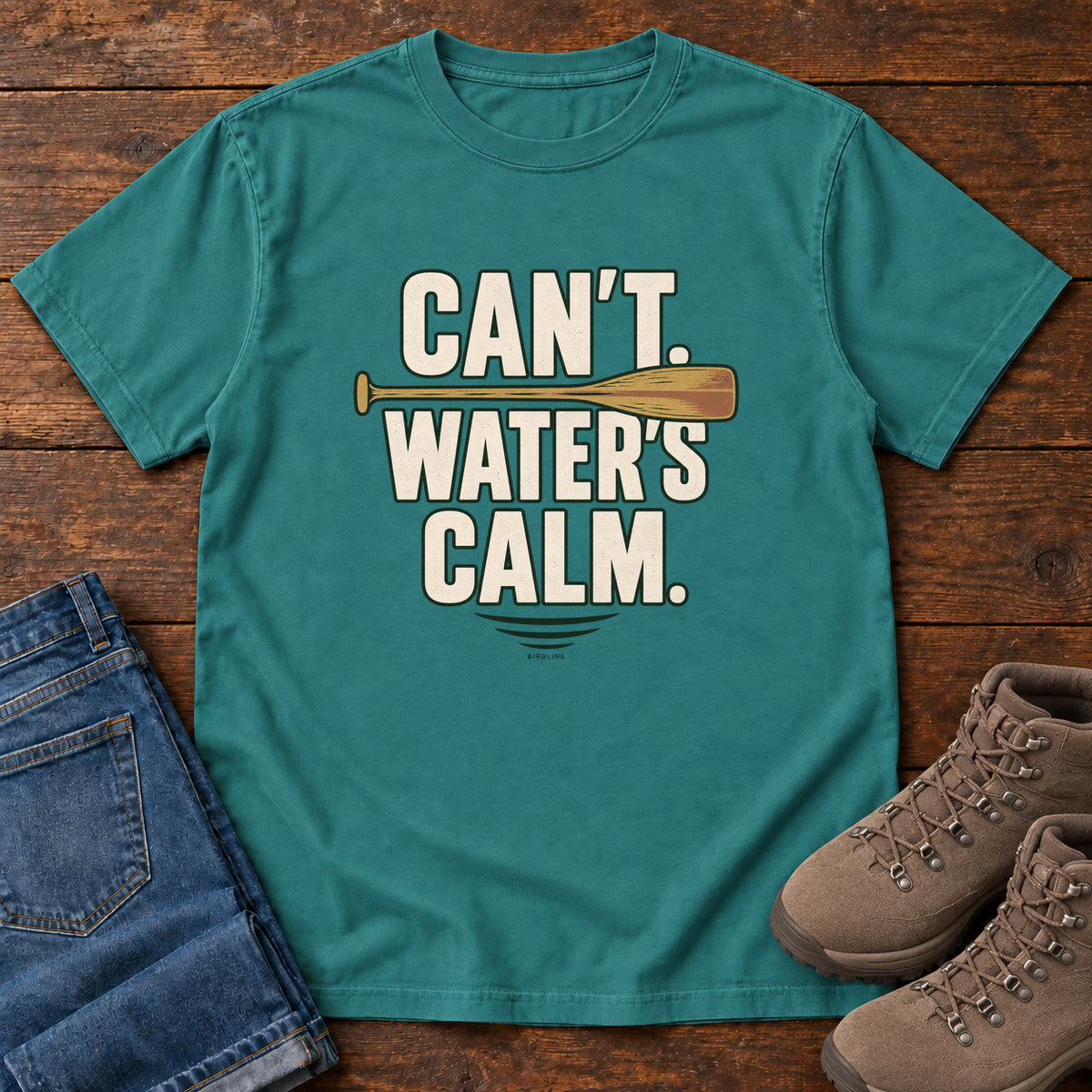 Can't. Water's Calm. T-Shirt in Emerald, front laid flat on wood, featuring bold cream slogan lettering divided by a horizontal orange paddle.