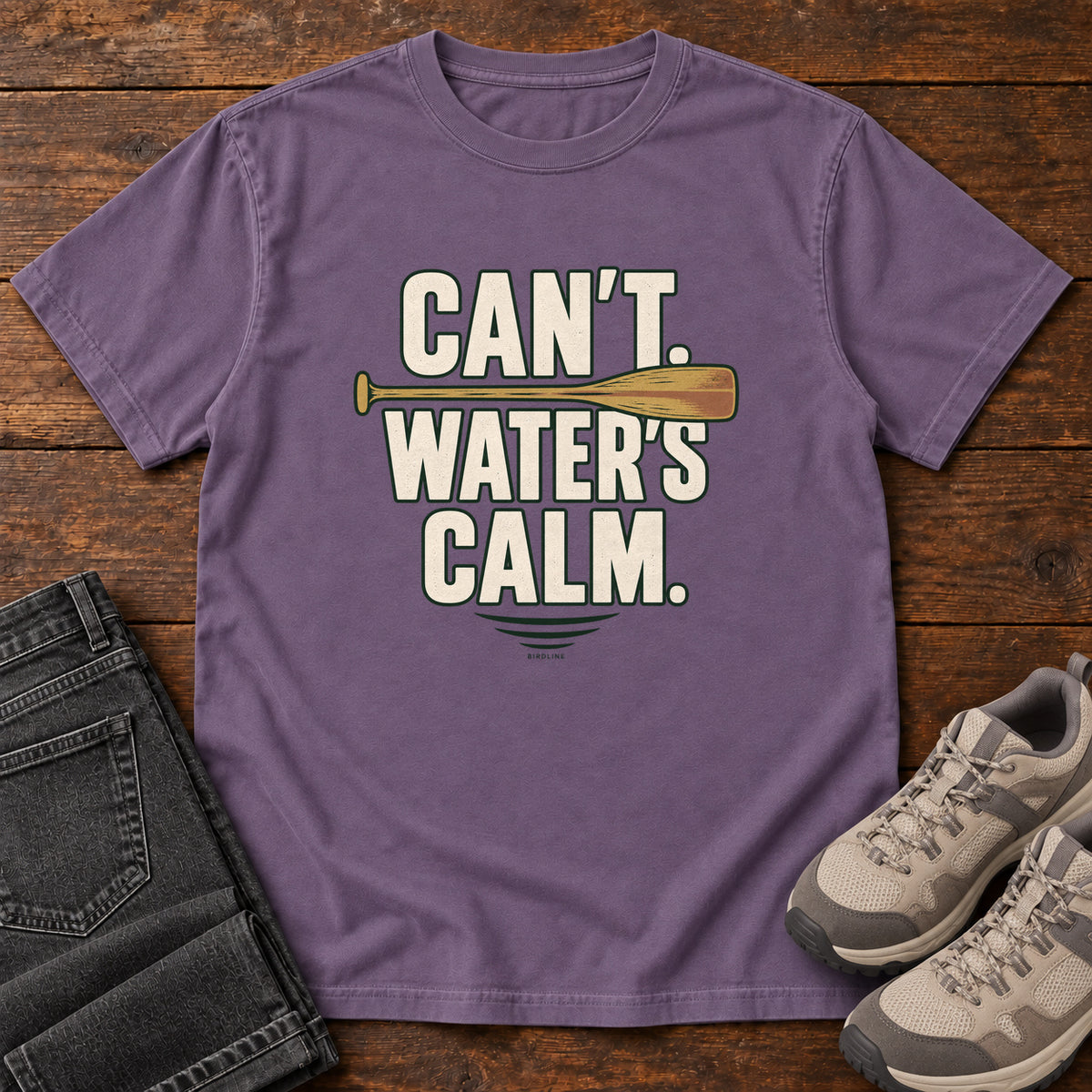 Can't. Water's Calm. T-Shirt in Grape, front laid flat on wood, featuring bold cream slogan lettering divided by a horizontal orange paddle.