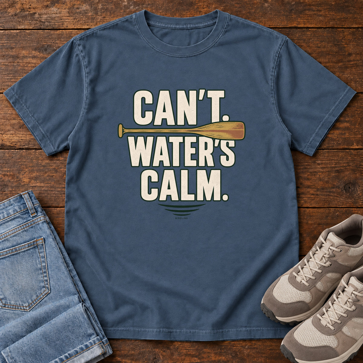 Can't. Water's Calm. T-Shirt in Midnight, front laid flat on wood, featuring bold cream slogan lettering divided by a horizontal orange paddle.