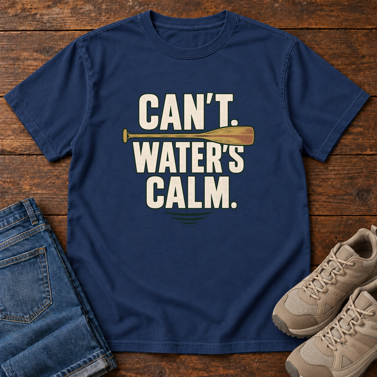 Can't. Water's Calm. T-Shirt in True Navy, front laid flat on wood, featuring bold cream slogan lettering divided by a horizontal orange paddle.