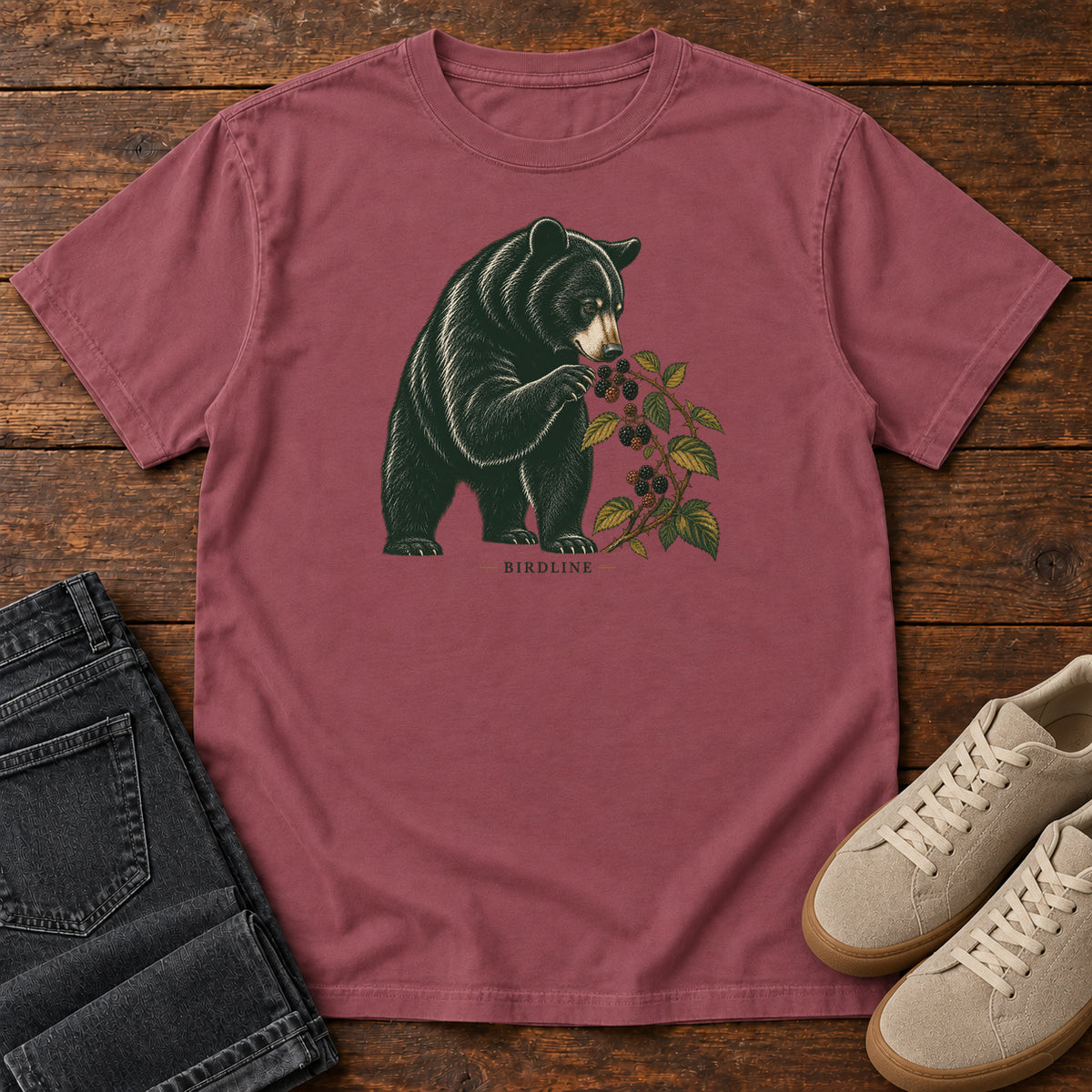 Brick Black Bear Berries Birdline T-Shirt with a dark bear holding a leafy berry branch, front flat lay on wood.