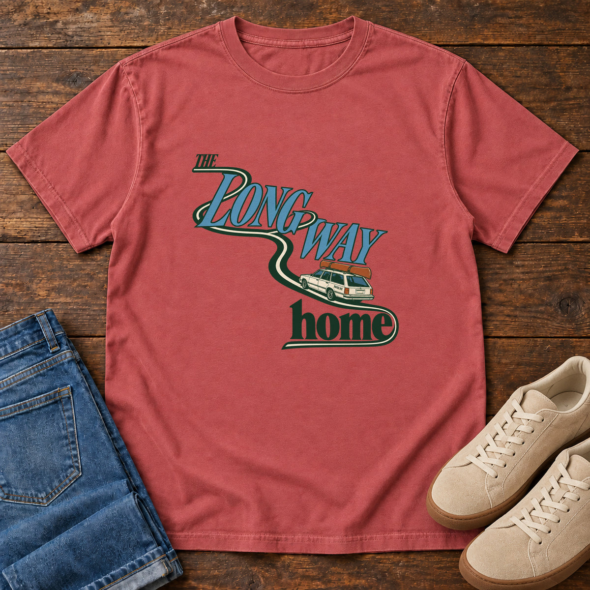 Crimson The Long Way Home T-Shirt with a station wagon on a winding road woven through The Long Way Home lettering; front flat lay on wood.