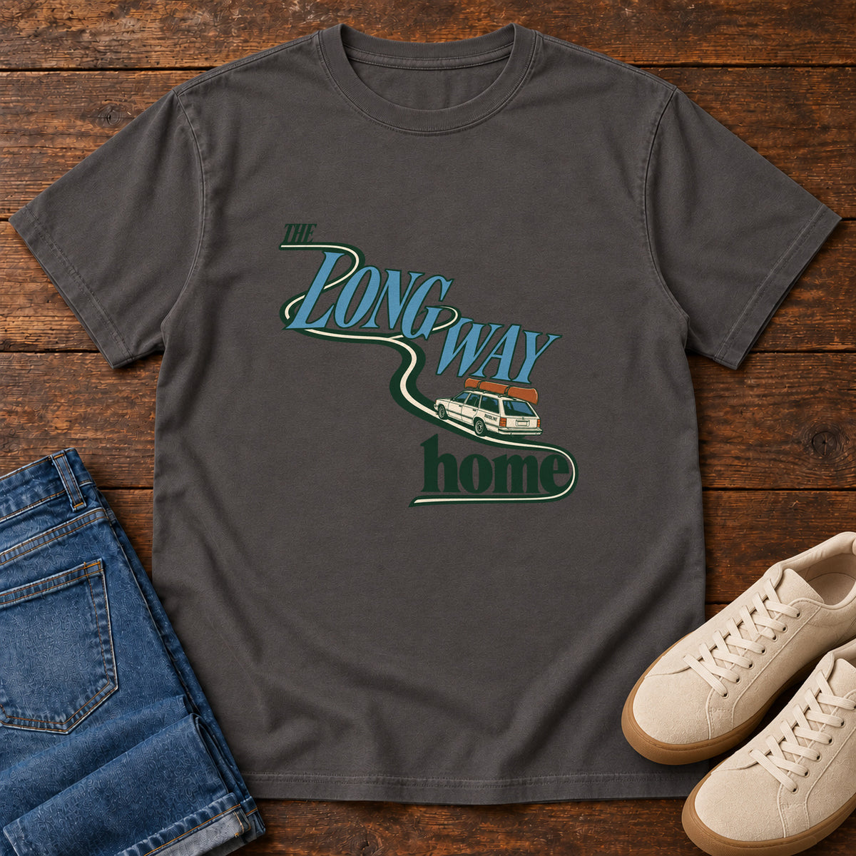 Graphite The Long Way Home T-Shirt with a station wagon on a winding road woven through The Long Way Home lettering; front flat lay on wood.