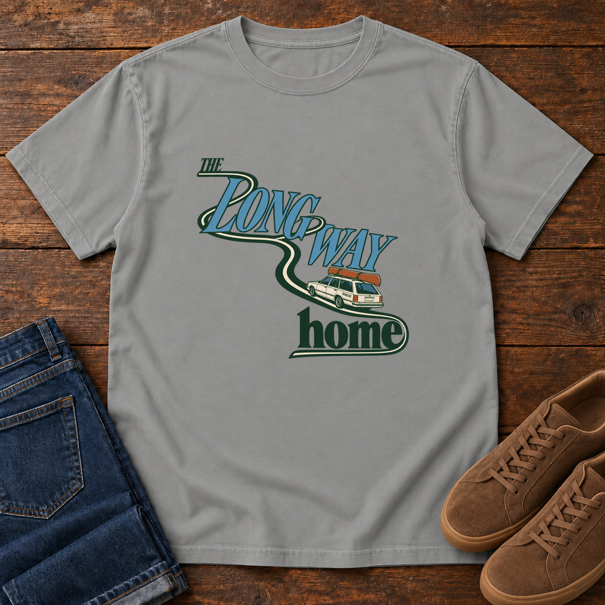 Grey The Long Way Home T-Shirt with a station wagon on a winding road woven through The Long Way Home lettering; front flat lay on wood.
