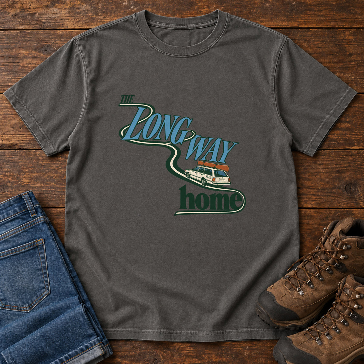 Pepper The Long Way Home T-Shirt with a station wagon on a winding road woven through The Long Way Home lettering; front flat lay on wood.