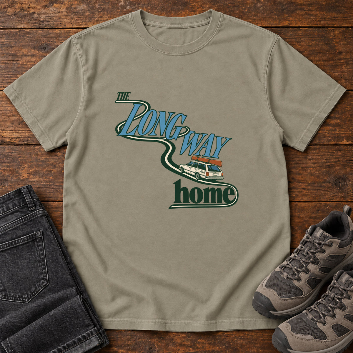 Sandstone The Long Way Home T-Shirt with a station wagon on a winding road woven through The Long Way Home lettering; front flat lay on wood.