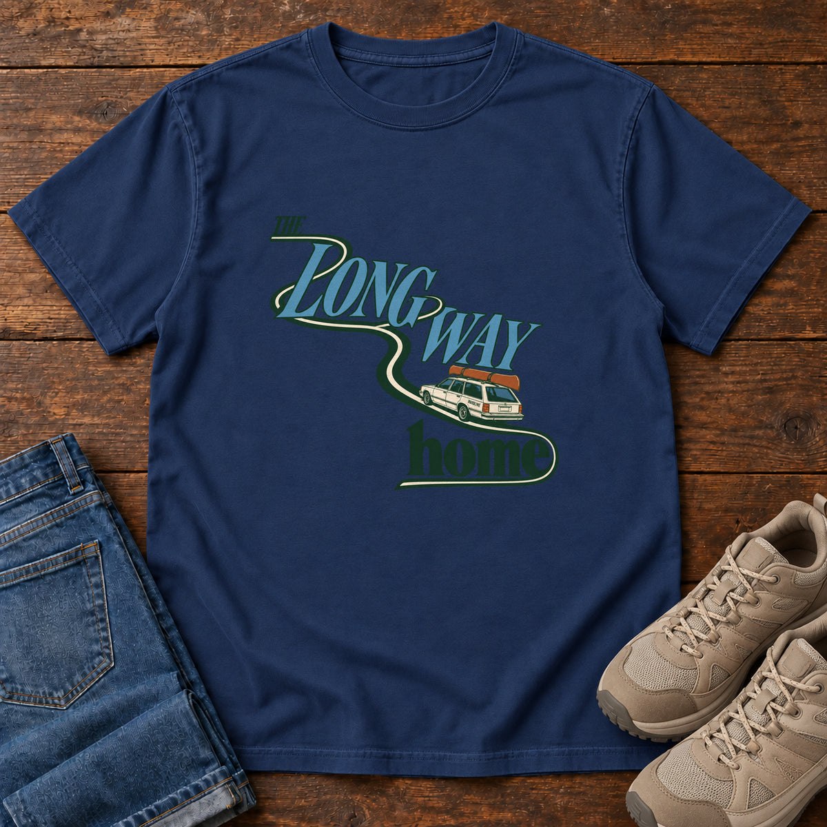True Navy The Long Way Home T-Shirt with a station wagon on a winding road woven through The Long Way Home lettering; front flat lay on wood.