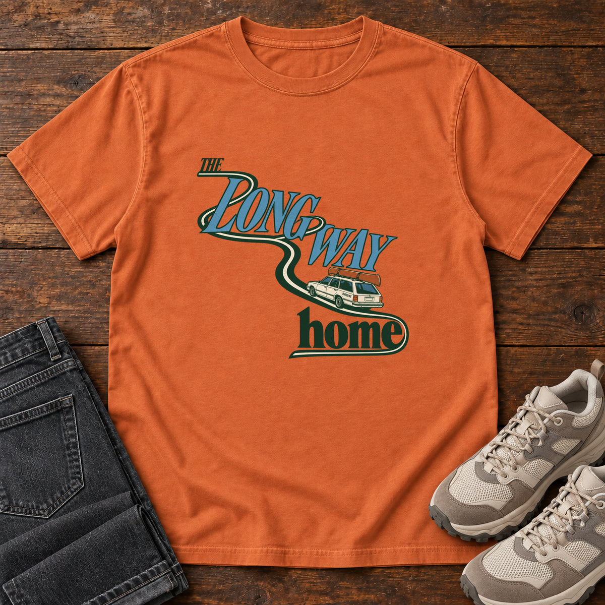 Yam The Long Way Home T-Shirt with a station wagon on a winding road woven through The Long Way Home lettering; front flat lay on wood.