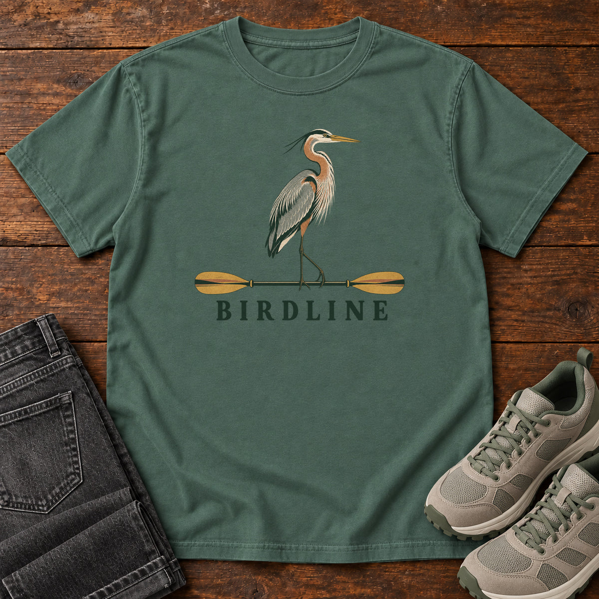 Blue Spruce Great Blue Heron Paddle Birdline T-Shirt with a long-legged heron standing on a horizontal paddle above Birdline lettering; front flat lay on wood.