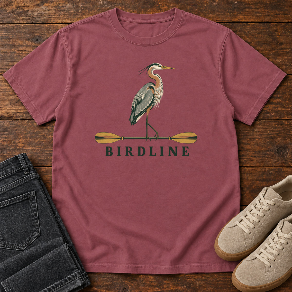 Brick Great Blue Heron Paddle Birdline T-Shirt with a long-legged heron standing on a horizontal paddle above Birdline lettering; front flat lay on wood.