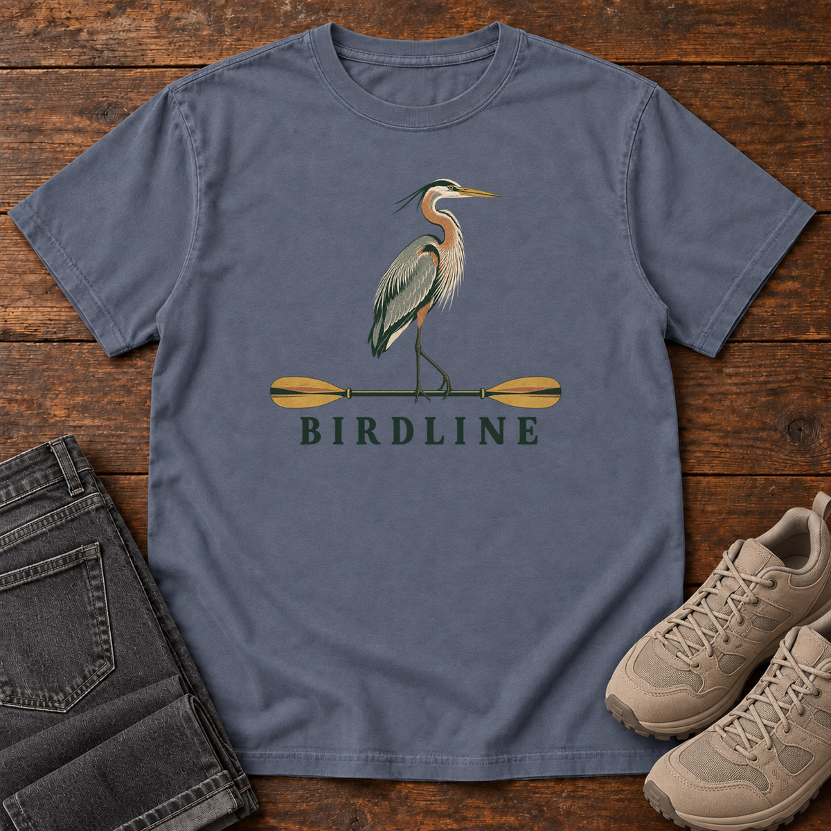 Denim Great Blue Heron Paddle Birdline T-Shirt with a long-legged heron standing on a horizontal paddle above Birdline lettering; front flat lay on wood.