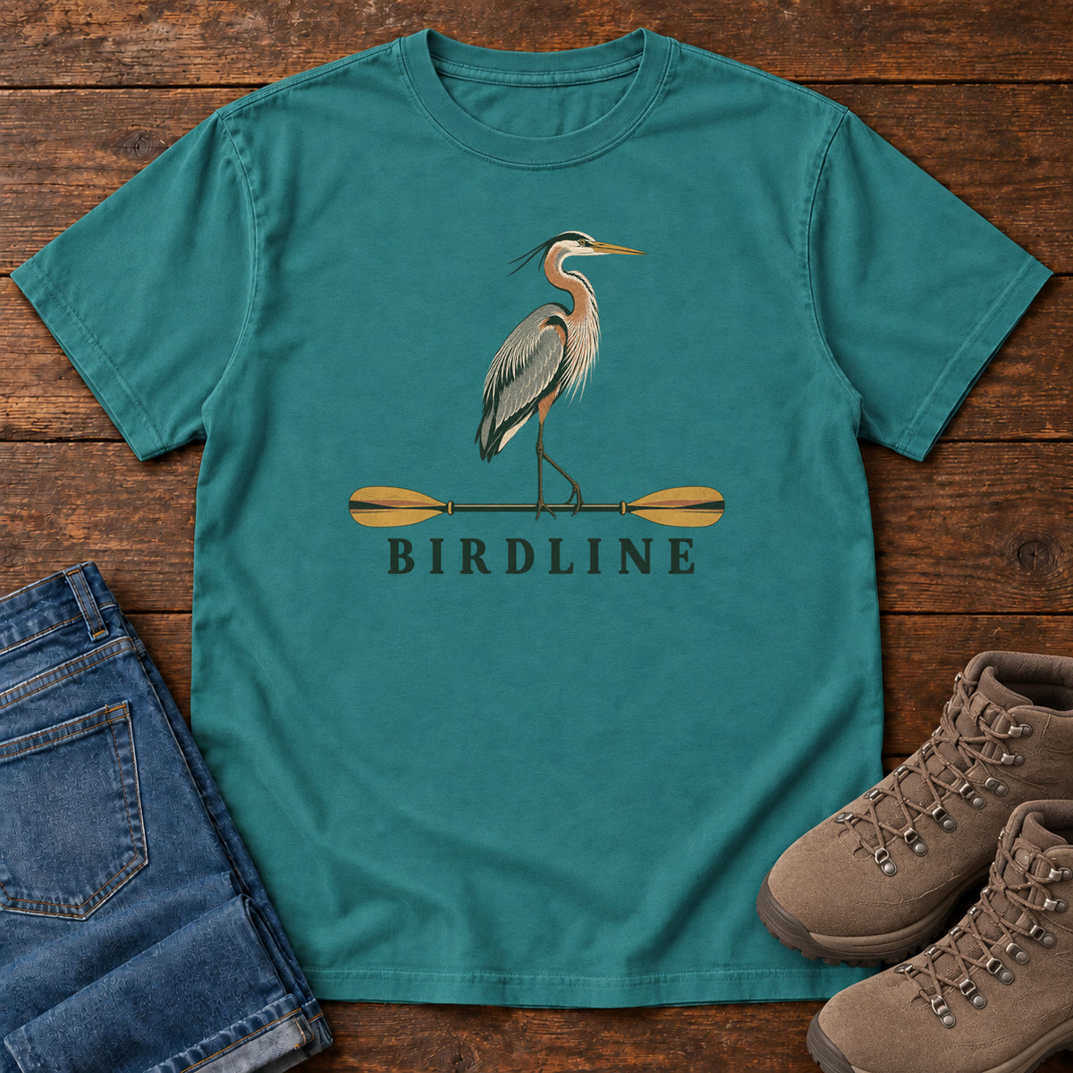 Emerald Great Blue Heron Paddle Birdline T-Shirt with a long-legged heron standing on a horizontal paddle above Birdline lettering; front flat lay on wood.