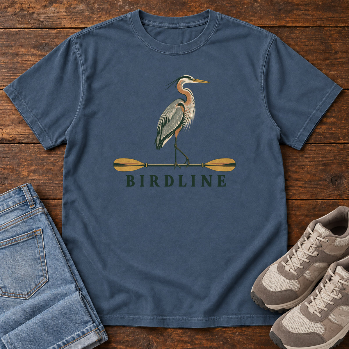 Midnight Great Blue Heron Paddle Birdline T-Shirt with a long-legged heron standing on a horizontal paddle above Birdline lettering; front flat lay on wood.