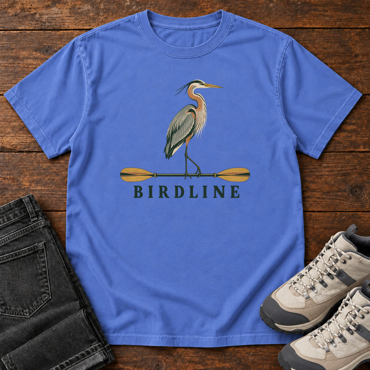 Mystic Blue Great Blue Heron Paddle Birdline T-Shirt with a long-legged heron standing on a horizontal paddle above Birdline lettering; front flat lay on wood.