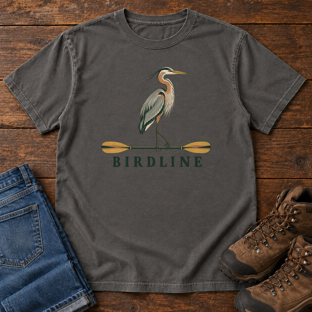 Pepper Great Blue Heron Paddle Birdline T-Shirt with a long-legged heron standing on a horizontal paddle above Birdline lettering; front flat lay on wood.