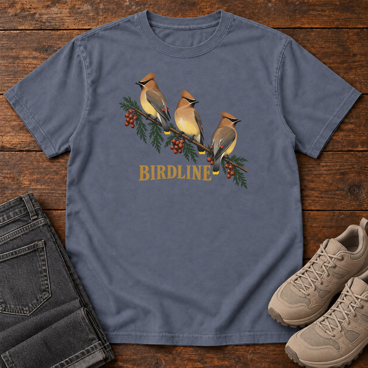 Denim Cedar Waxwings Birdline T-Shirt with three crested birds on a berry-laden evergreen branch; front flat lay on wood.
