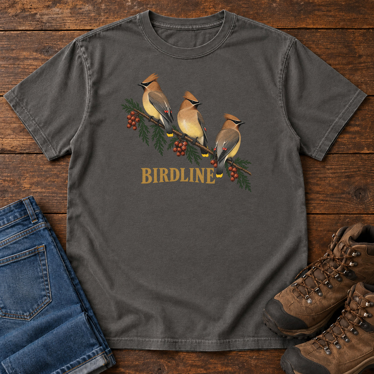 Pepper Cedar Waxwings Birdline T-Shirt with three crested birds on a berry-laden evergreen branch; front flat lay on wood.