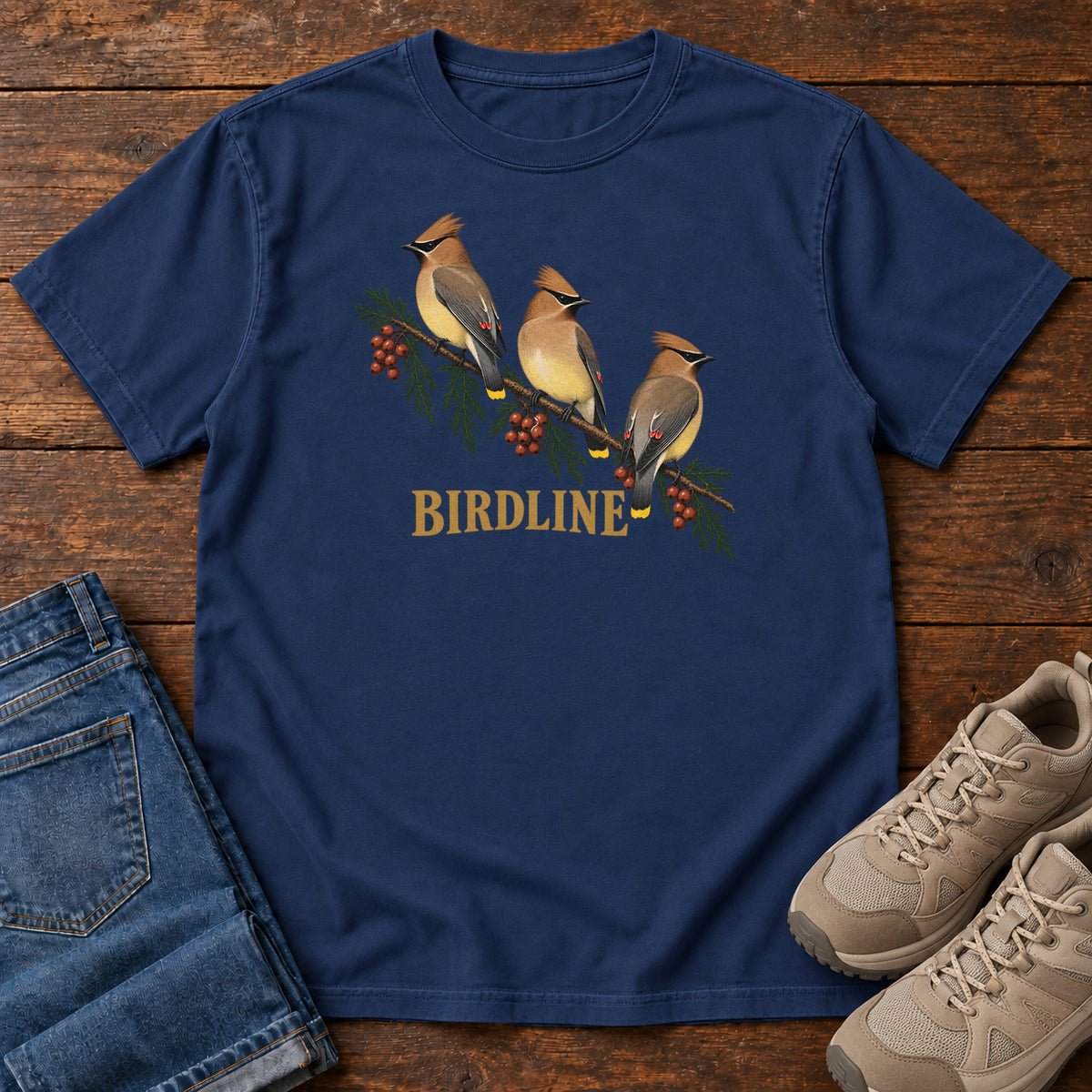 True Navy Cedar Waxwings Birdline T-Shirt with three crested birds on a berry-laden evergreen branch; front flat lay on wood.