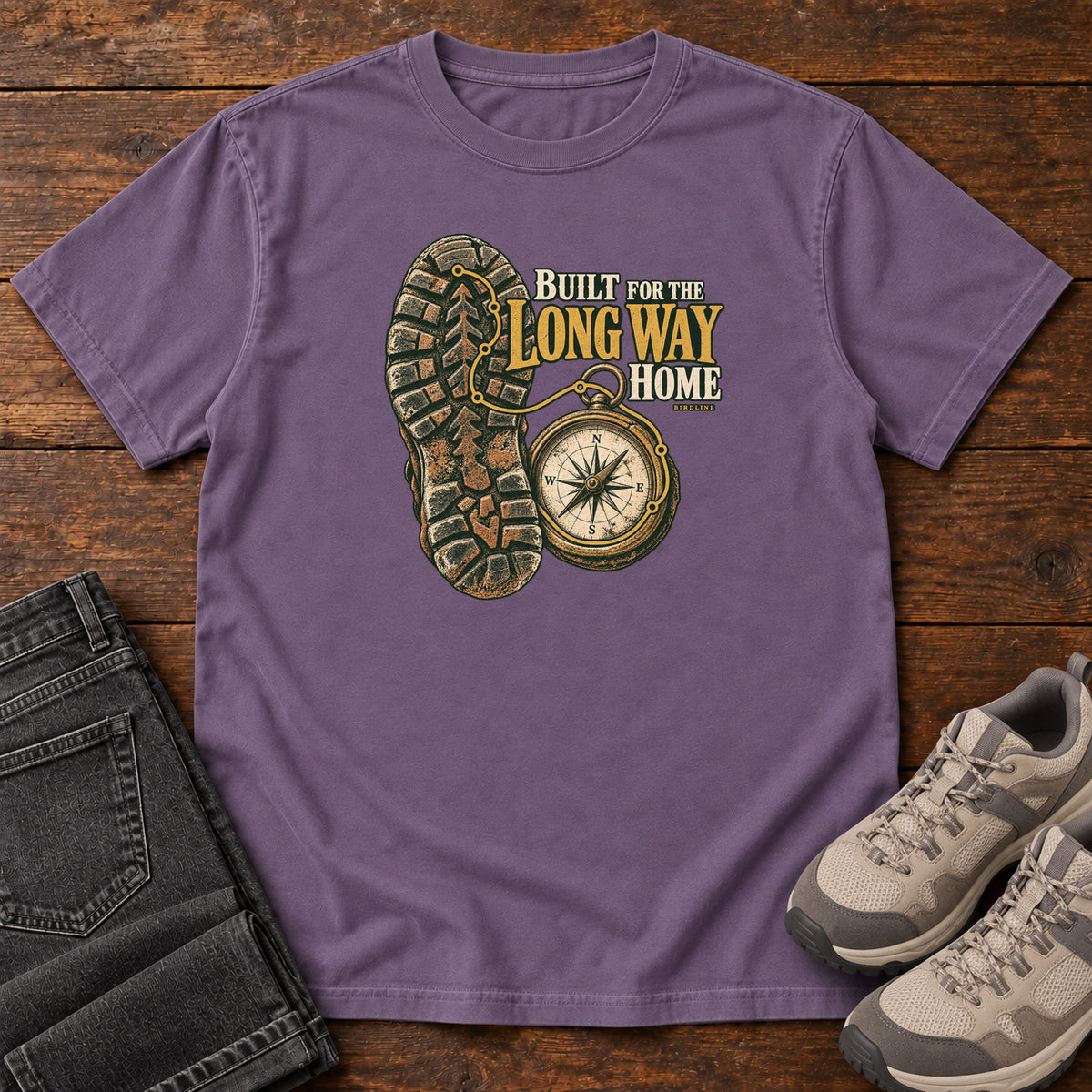 Built for the Long Way Home T-Shirt in Grape, front laid flat on wood, featuring a rugged boot sole beside a round compass and cream-and-gold slogan lettering.