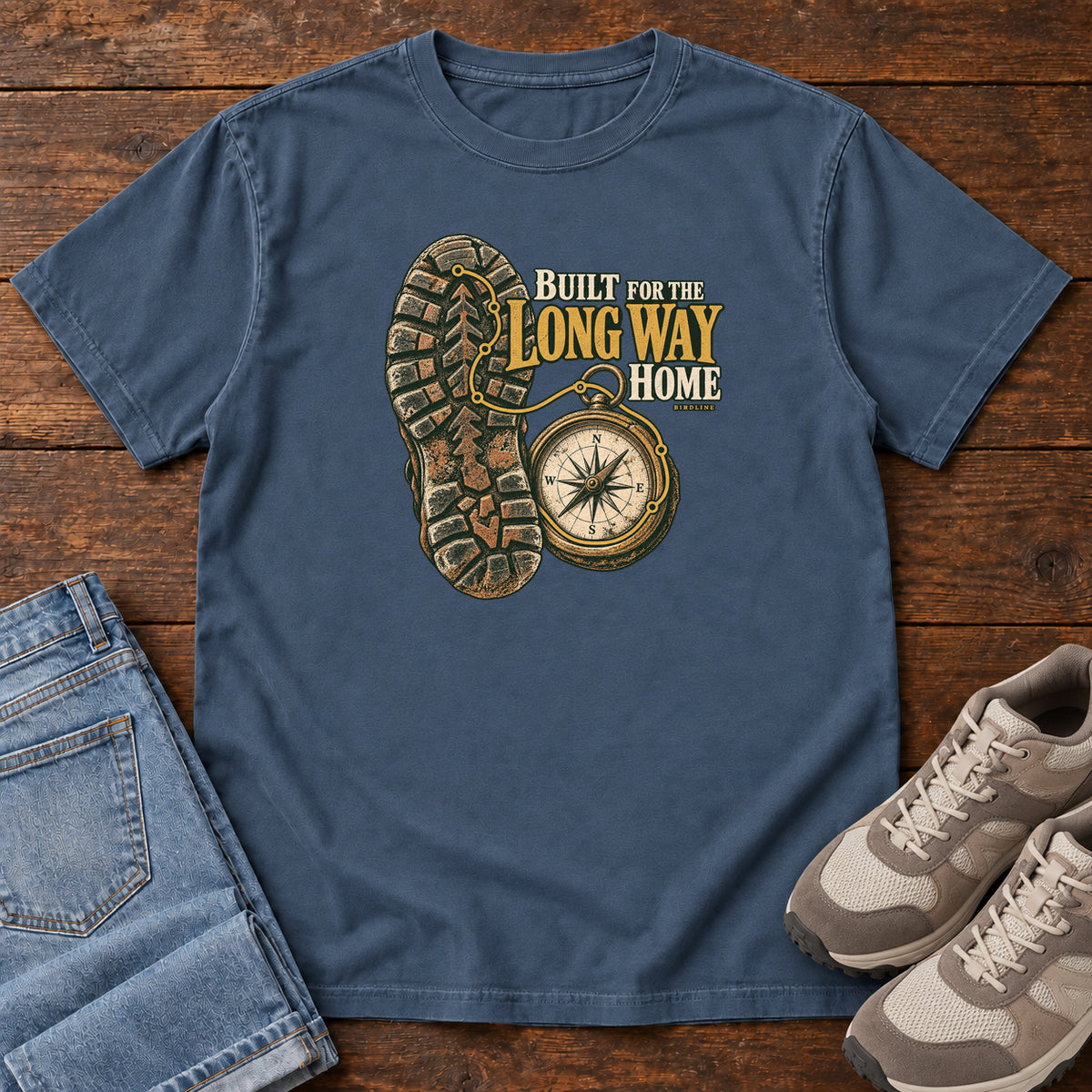 Built for the Long Way Home T-Shirt in Midnight, front laid flat on wood, featuring a rugged boot sole beside a round compass and cream-and-gold slogan lettering.