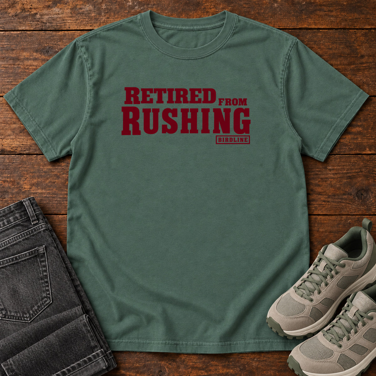 Blue Spruce Retired From Rushing T-Shirt with burgundy Retired From Rushing lettering; front flat lay on wood.