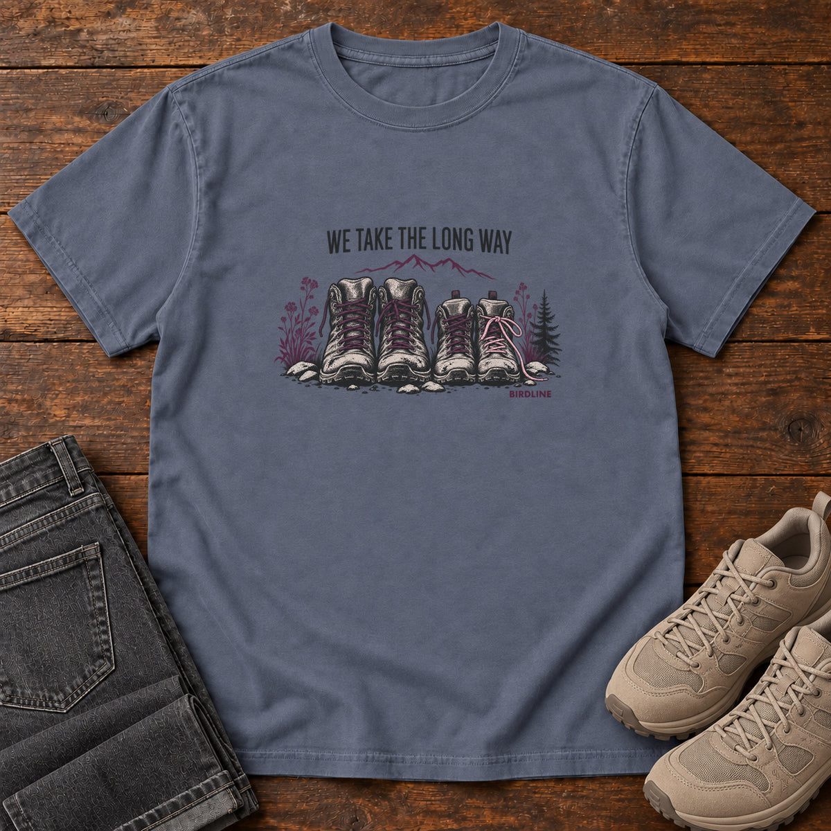 We Take the Long Way T-Shirt in Denim, front laid flat on wood, featuring two pairs of hiking boots with stones, foliage and a mountain outline beneath slogan lettering.