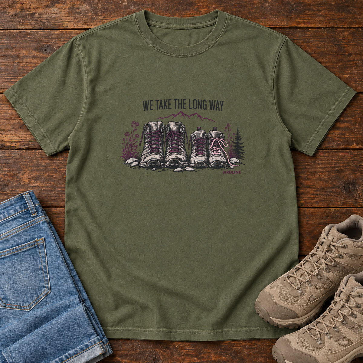 We Take the Long Way T-Shirt in Hemp, front laid flat on wood, featuring two pairs of hiking boots with stones, foliage and a mountain outline beneath slogan lettering.