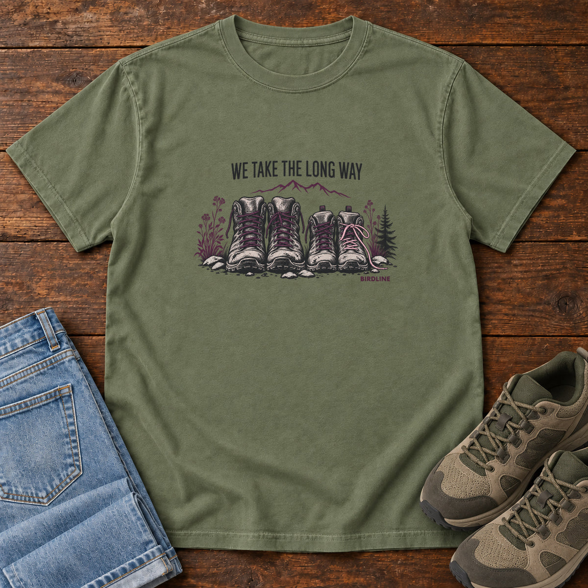 We Take the Long Way T-Shirt in Moss, front laid flat on wood, featuring two pairs of hiking boots with stones, foliage and a mountain outline beneath slogan lettering.