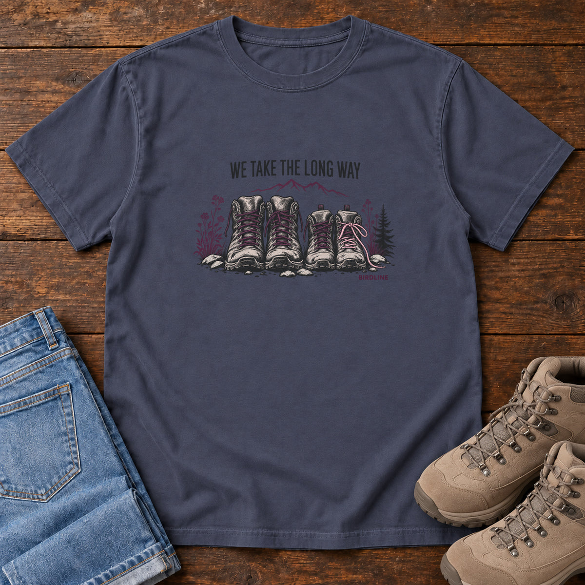 We Take the Long Way T-Shirt in Navy, front laid flat on wood, featuring two pairs of hiking boots with stones, foliage and a mountain outline beneath slogan lettering.