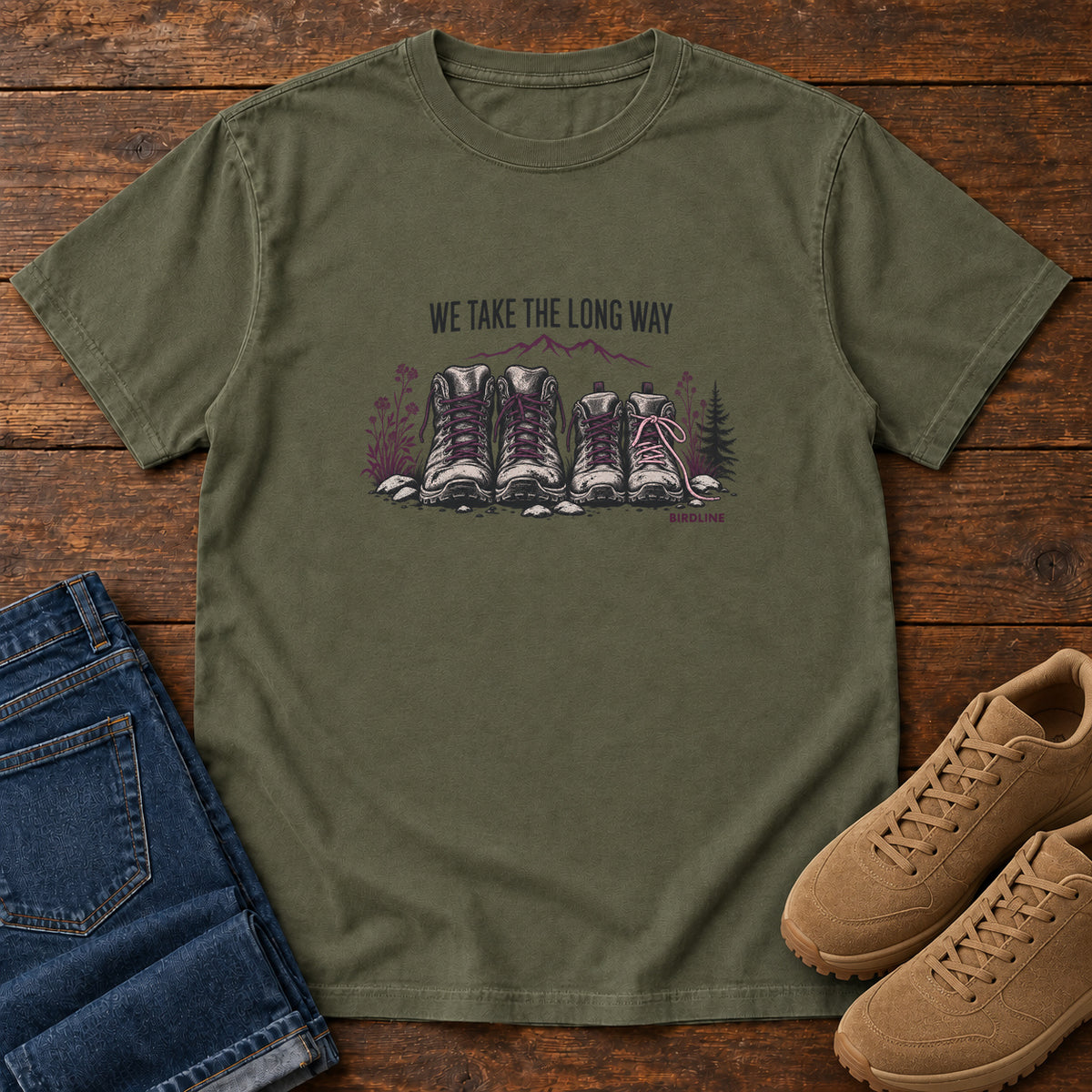 We Take the Long Way T-Shirt in Sage, front laid flat on wood, featuring two pairs of hiking boots with stones, foliage and a mountain outline beneath slogan lettering.