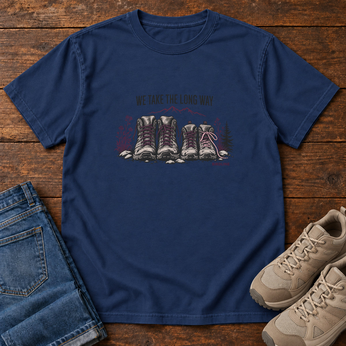 We Take the Long Way T-Shirt in True Navy, front laid flat on wood, featuring two pairs of hiking boots with stones, foliage and a mountain outline beneath slogan lettering.