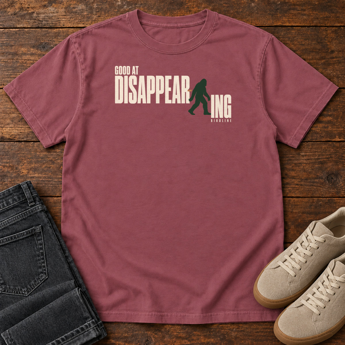 Brick Good at Disappearing T-Shirt with cream lettering interrupted by a green walking Bigfoot silhouette; front flat lay on wood.