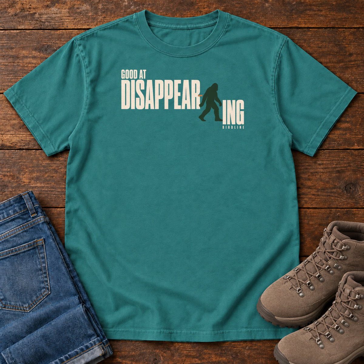 Emerald Good at Disappearing T-Shirt with cream lettering interrupted by a green walking Bigfoot silhouette; front flat lay on wood.
