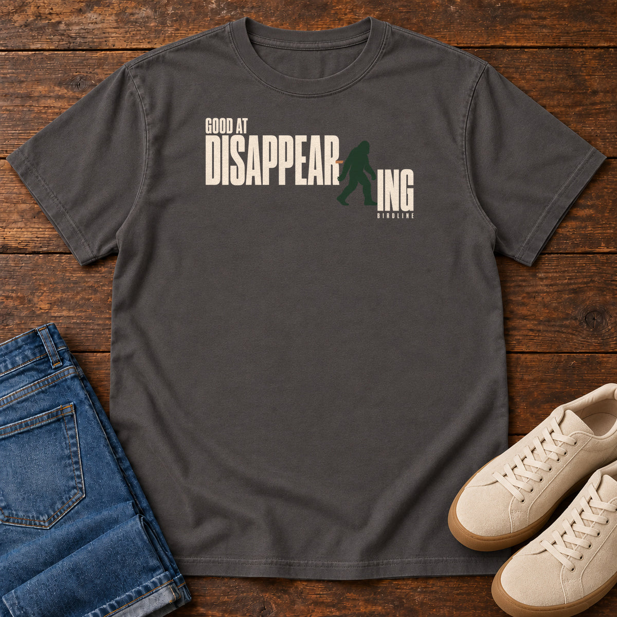 Graphite Good at Disappearing T-Shirt with cream lettering interrupted by a green walking Bigfoot silhouette; front flat lay on wood.