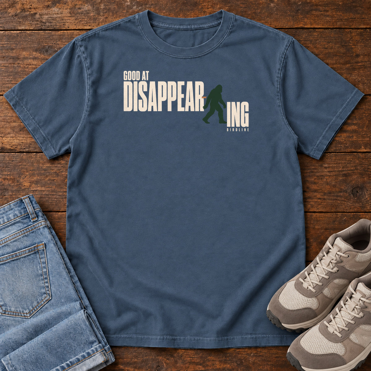 Midnight Good at Disappearing T-Shirt with cream lettering interrupted by a green walking Bigfoot silhouette; front flat lay on wood.