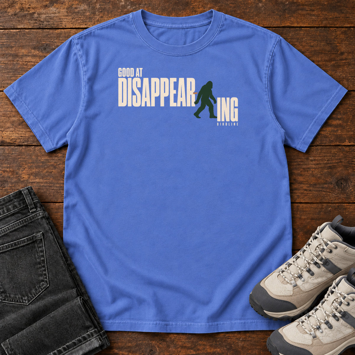 Mystic Blue Good at Disappearing T-Shirt with cream lettering interrupted by a green walking Bigfoot silhouette; front flat lay on wood.