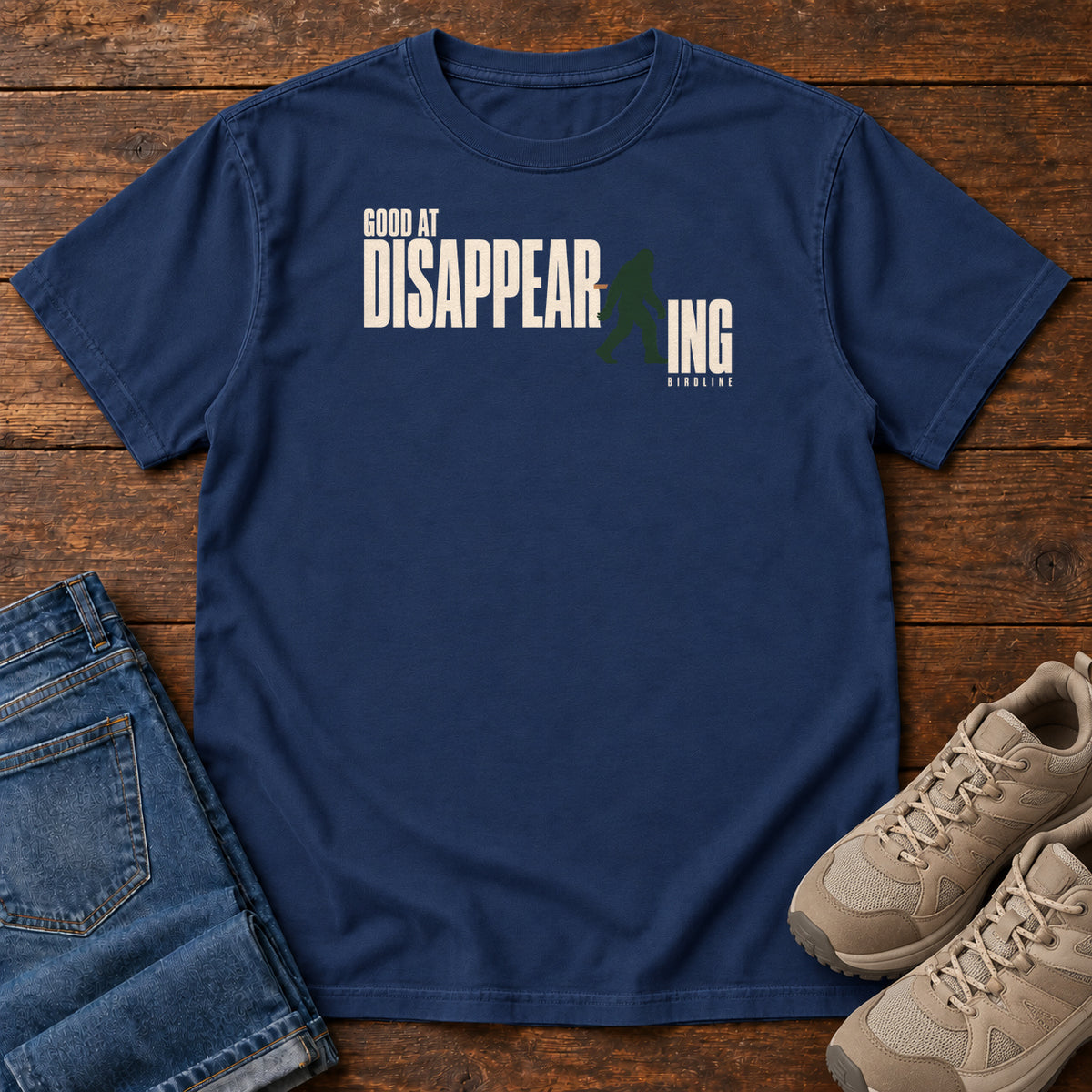 True Navy Good at Disappearing T-Shirt with cream lettering interrupted by a green walking Bigfoot silhouette; front flat lay on wood.