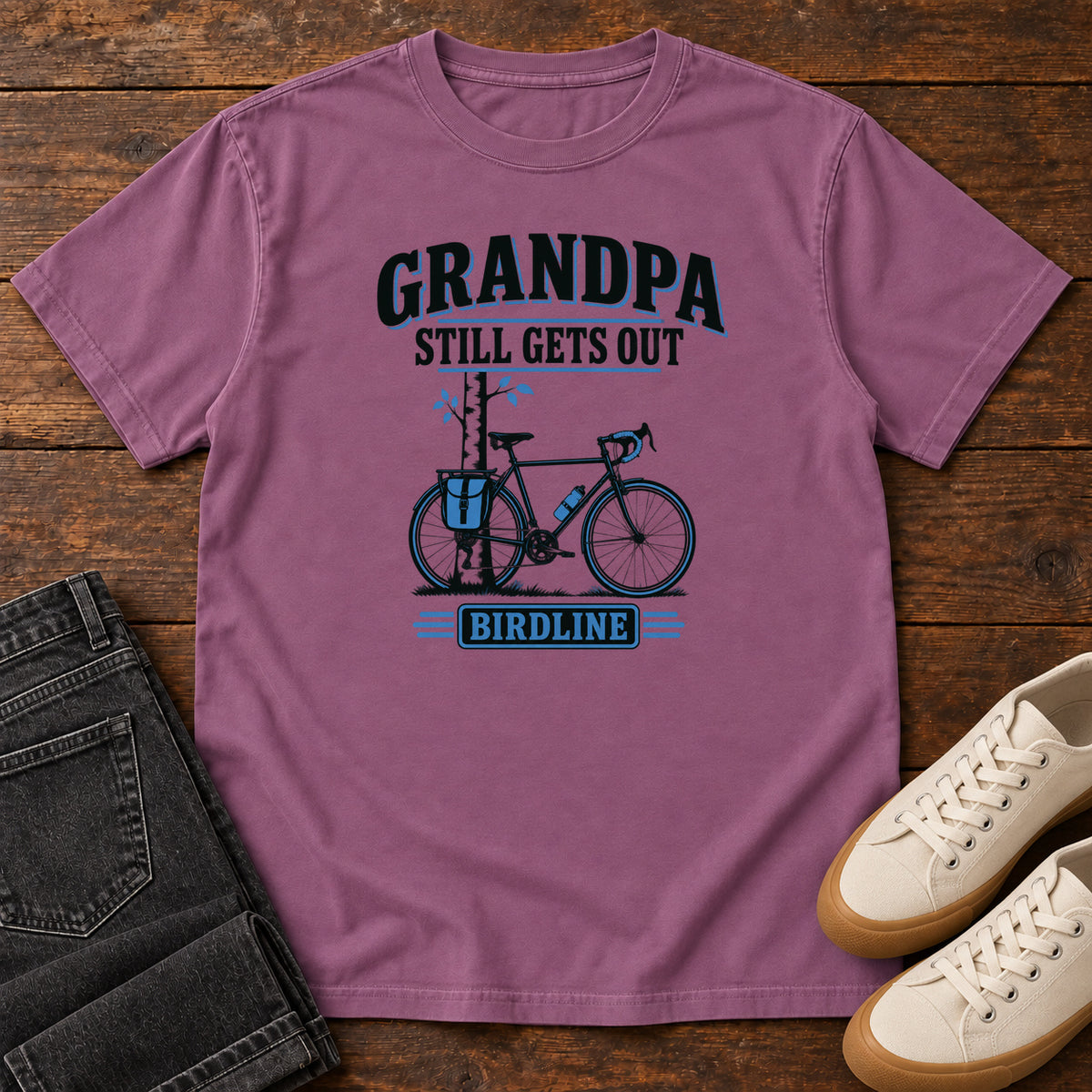 Berry Grandpa Still Gets Out T-Shirt with a bicycle beside a slender tree and blue-accented lettering, front flat lay on wood.