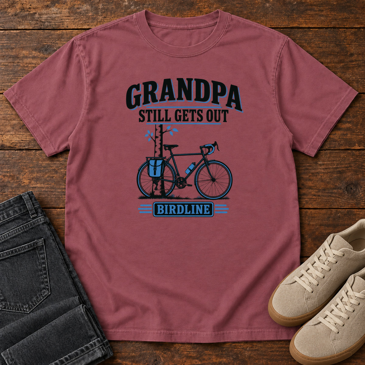 Brick Grandpa Still Gets Out T-Shirt with a bicycle beside a slender tree and blue-accented lettering, front flat lay on wood.
