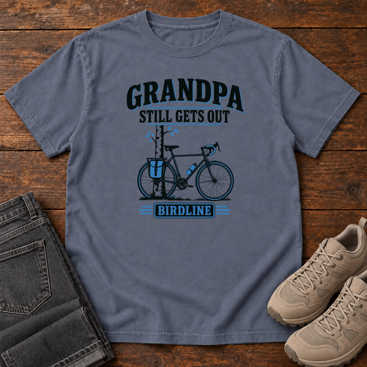 Denim Grandpa Still Gets Out T-Shirt with a bicycle beside a slender tree and blue-accented lettering, front flat lay on wood.