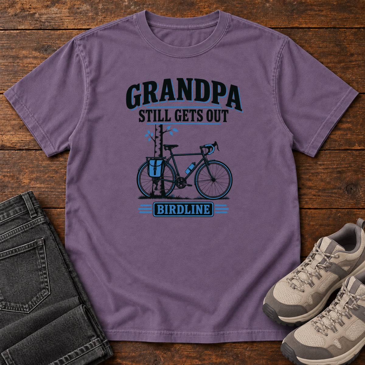 Grape Grandpa Still Gets Out T-Shirt with a bicycle beside a slender tree and blue-accented lettering, front flat lay on wood.