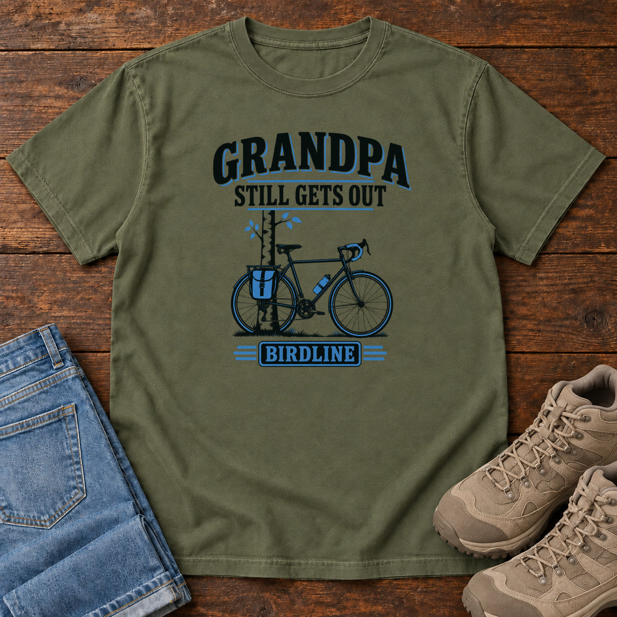 Hemp Grandpa Still Gets Out T-Shirt with a bicycle beside a slender tree and blue-accented lettering, front flat lay on wood.
